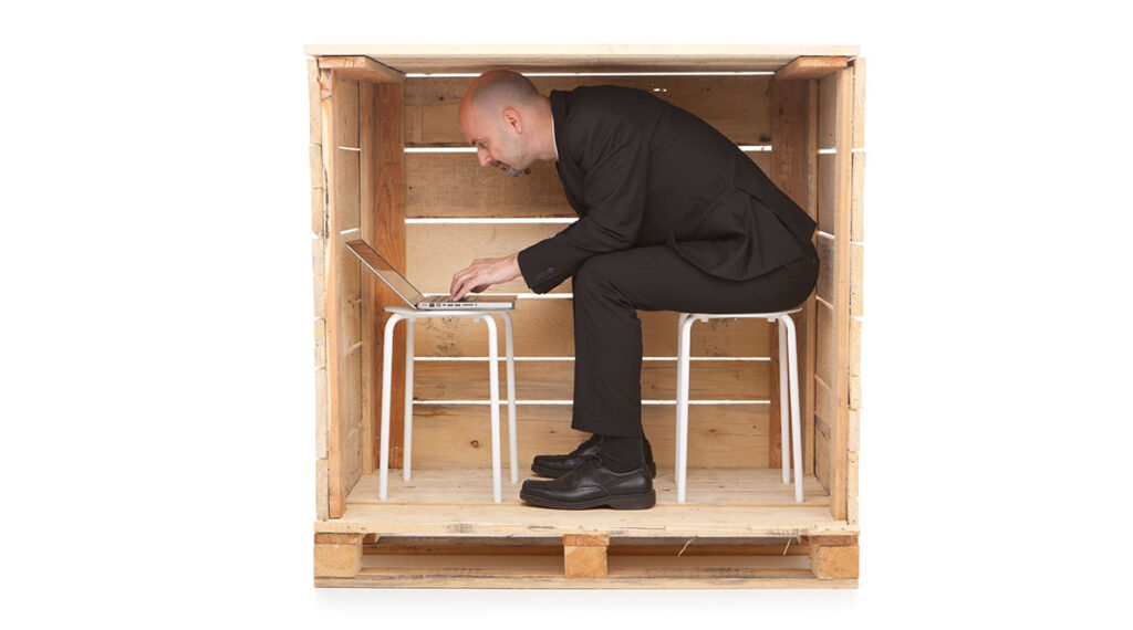 A man in a wooden box hunched over working on a laptop