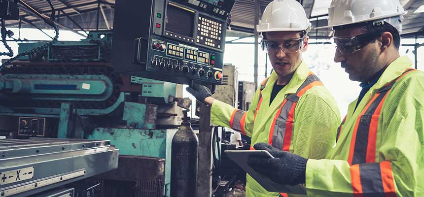 Two men in high visibility vests and hardhats are looking at a tablet device in from of machinery