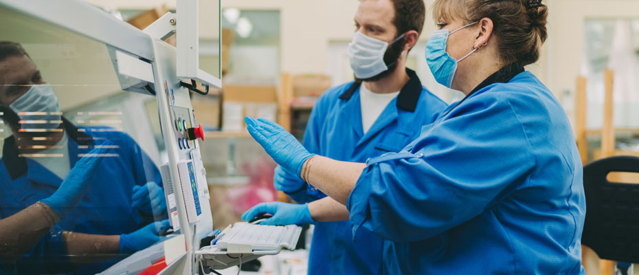 Two people in lab coats and face masks work a piece of machinery