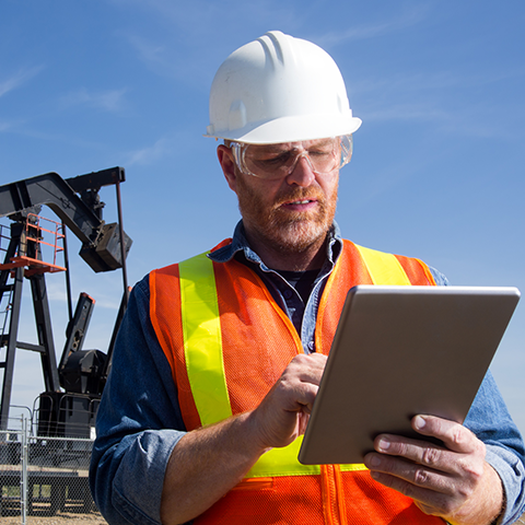 A man standing in front of an oil rig wearing a hard hat and using a tablet device.