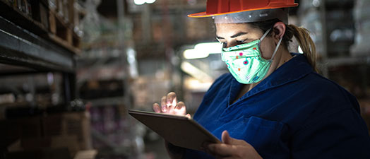 A woman in a facemask and a hardhat working in a warehouse using a tablet device.